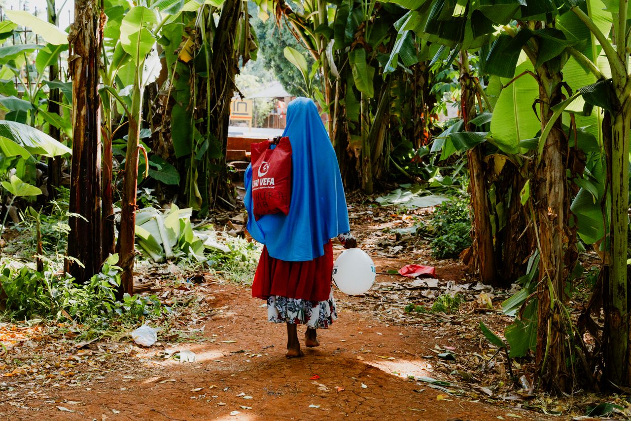 A woman in vibrant clothing walks along a jungle path, carrying a ball and a bag.
