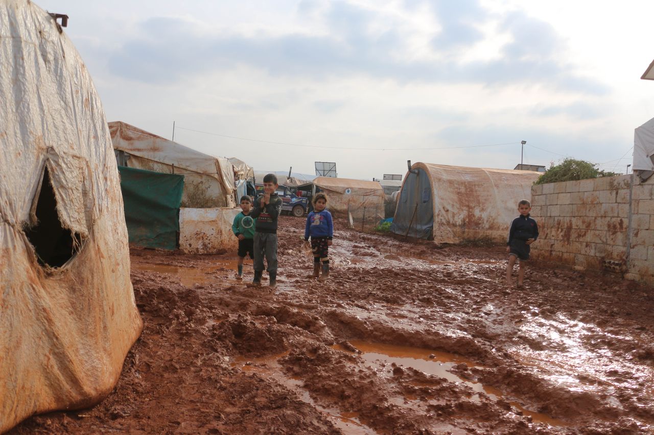 Distant carefree ethnic children standing on dirty ground near old tents in poor district of refugee camp in small settlement